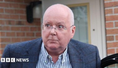 A bespectacled balding man in a blue sports jacket and striped shirt emerges from a car outside the front door of a modern house