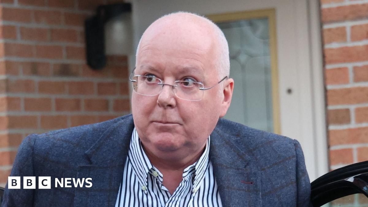 A bespectacled balding man in a blue sports jacket and striped shirt emerges from a car outside the front door of a modern house