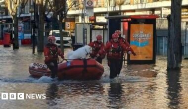 A street lines with houses and flooded. A boat is being led down the road by three men in high viz. A dog is in the boat.