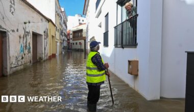 A woman looks down from a balcony on a man standing in a flooded street.