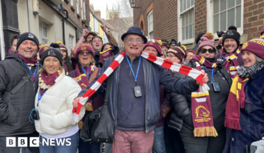 The picture shows more than a dozen men and women smiling at the camera. The majority are wearing burgundy bobble hats, and matching scarves. The man in the middle is wearing a red and white striped scarf around his neck, which he is holding out with his arms. They are standing in front of a row of houses.