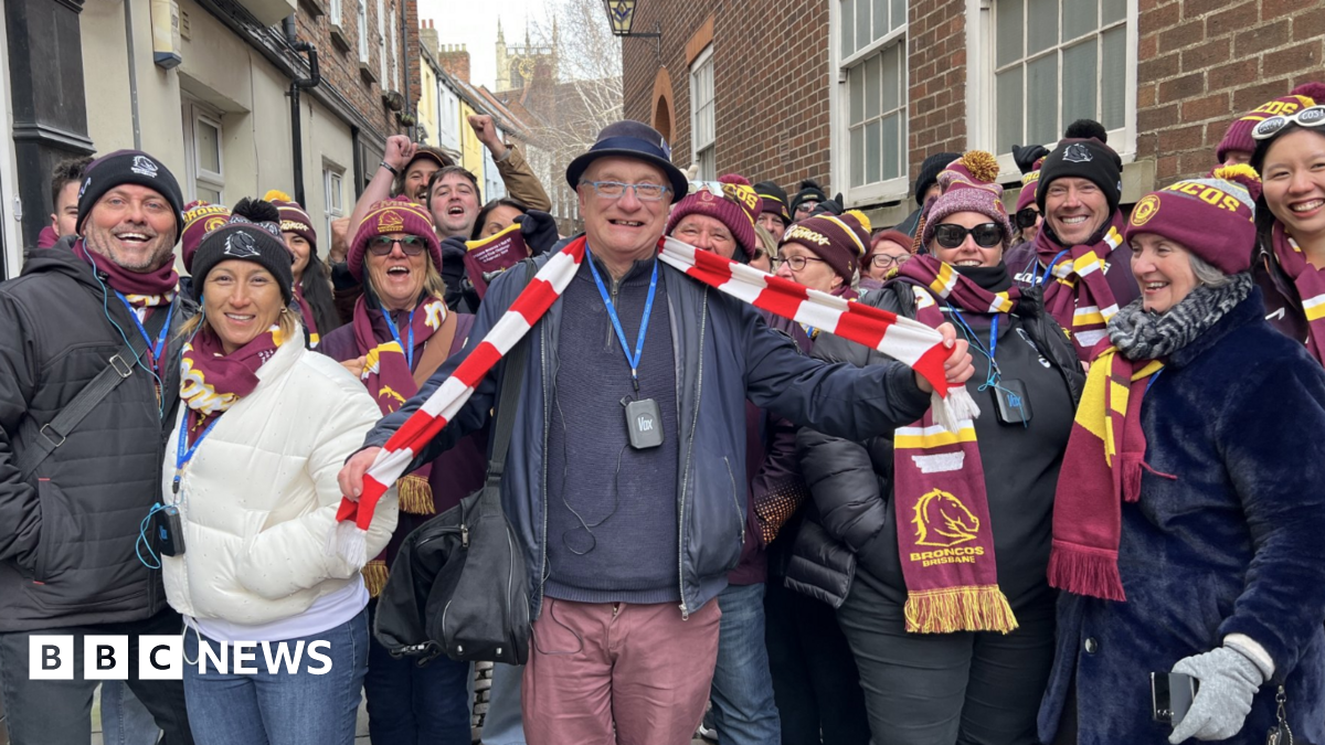 The picture shows more than a dozen men and women smiling at the camera. The majority are wearing burgundy bobble hats, and matching scarves. The man in the middle is wearing a red and white striped scarf around his neck, which he is holding out with his arms. They are standing in front of a row of houses.