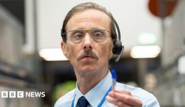 Mackenzie Crook as a shop worker with headset and glasses. he wears a shirt and tie and points with a pen.