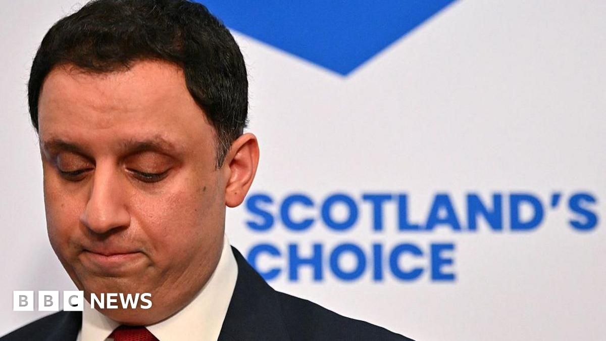 Anas Sarwar in dark suit and white shirt with red tie looks down as though in thought. A blue and white saltire backdrop with the words "Scotland's Choice" is directly behind him.