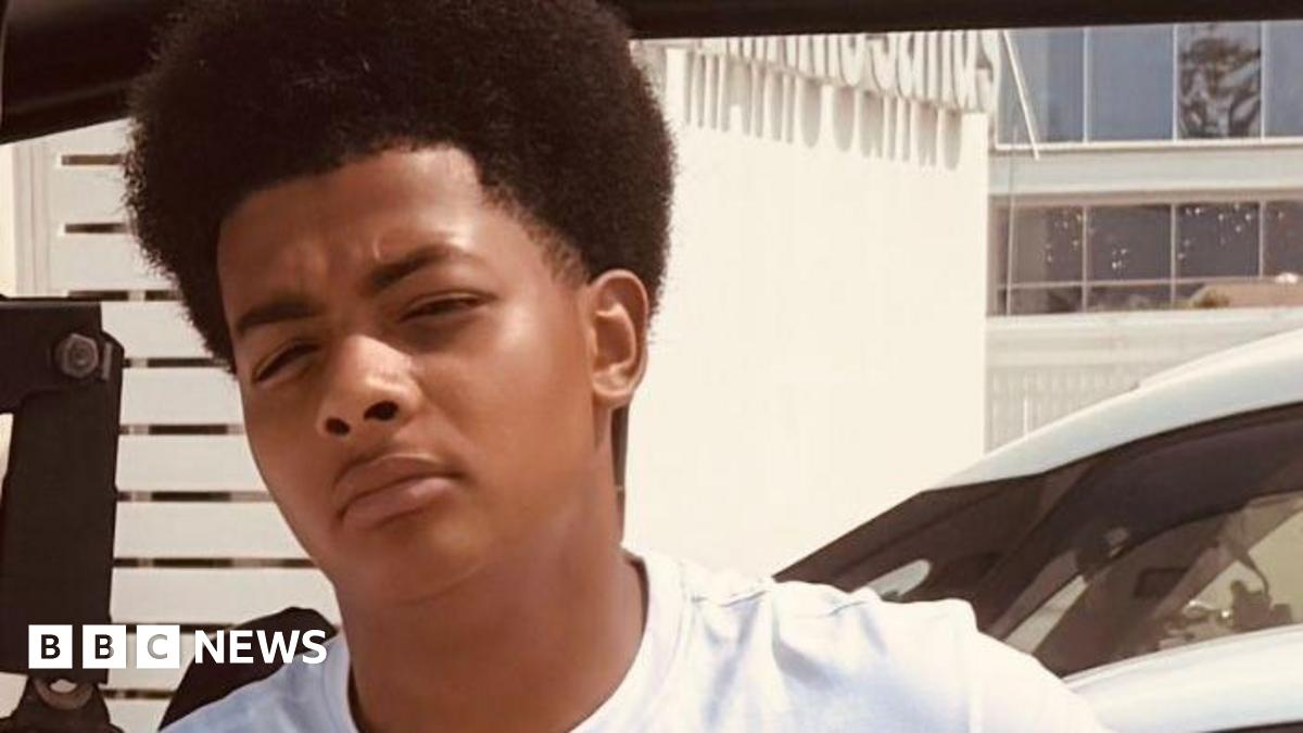 A teenage boy leaning against a car, wearing a white top