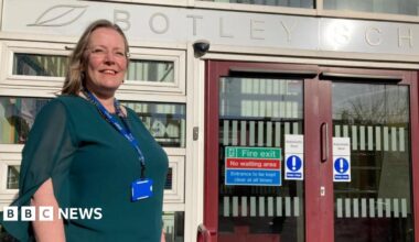 A woman is standing outside a school entrance. She is wearing glasses and a green top and looks very proud.