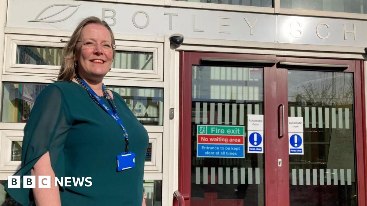 A woman is standing outside a school entrance. She is wearing glasses and a green top and looks very proud.