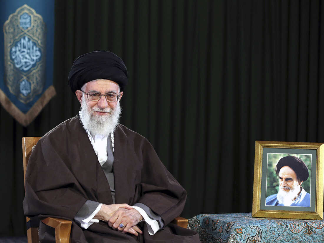 In this 2017 photo, Ayatollah Ali Khamenei, wearing a turban and traditional clothing, sits on a chair. A framed portrait of Ayatollah Ruhollah Khomeini stands on a table on the right side of the frame.