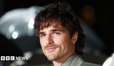 Jacob Elordi holds a phone up to take a selfie with two fans, one of whom is holding an umbrella, at the Wuthering Heights premiere. Elordi has dark hair and a moustache, and is wearing a grey suit.