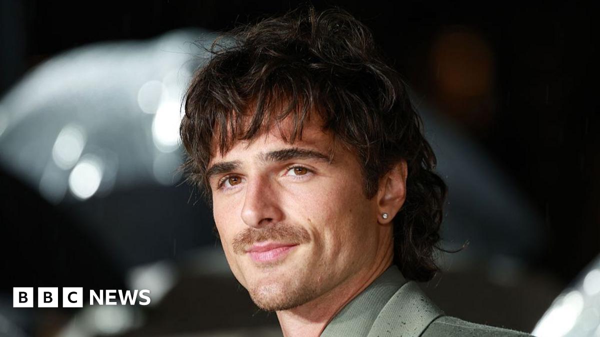 Jacob Elordi holds a phone up to take a selfie with two fans, one of whom is holding an umbrella, at the Wuthering Heights premiere. Elordi has dark hair and a moustache, and is wearing a grey suit.