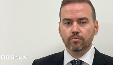 A man with short, light brown hair, wears a blue suit, white shirt and blue polka dot tie. He has a short bear and stands against a plain white background with a neutral expression on his face.