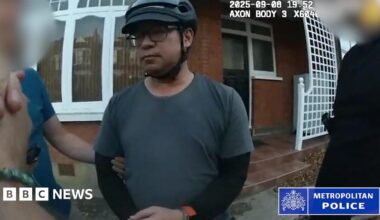 A still from bodycam footage of the police arrest of Vincent Chan. He is outside a house and is weaing a grey T-shirt with a black long-sleeved top underneath. He is wearing glasses and a bicycle helmet. Police are standing either side of him and his arm is being held.
