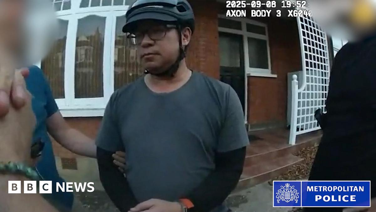 A still from bodycam footage of the police arrest of Vincent Chan. He is outside a house and is weaing a grey T-shirt with a black long-sleeved top underneath. He is wearing glasses and a bicycle helmet. Police are standing either side of him and his arm is being held.