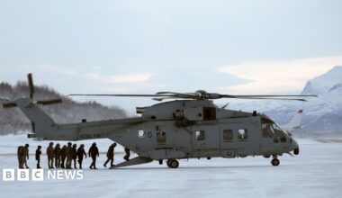 A group of people board a grey army helicopter in Norway with snowy mountains in the background