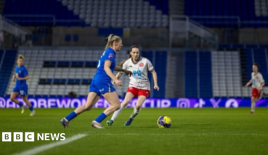 Players from Birmingham City and Chatham Town are on the pitch, with other team members in the background. There are football stands behind them.