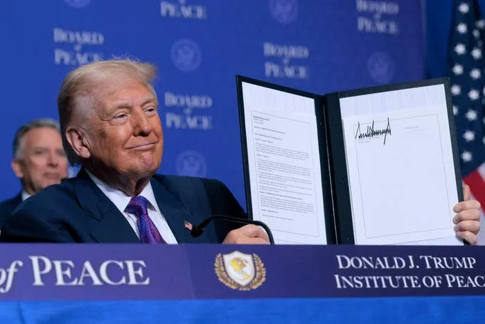 A person in a suit holds up a signed document at an event labeled "Donald J. Trump Institute of Peace."