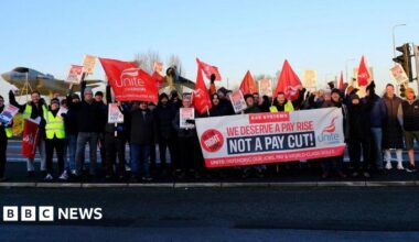 A small group of protesters holding flags and placards pushing for a pay rise. They are stood at the side of a road and dressed warmly in puffer jackets, jeans and woolly hats