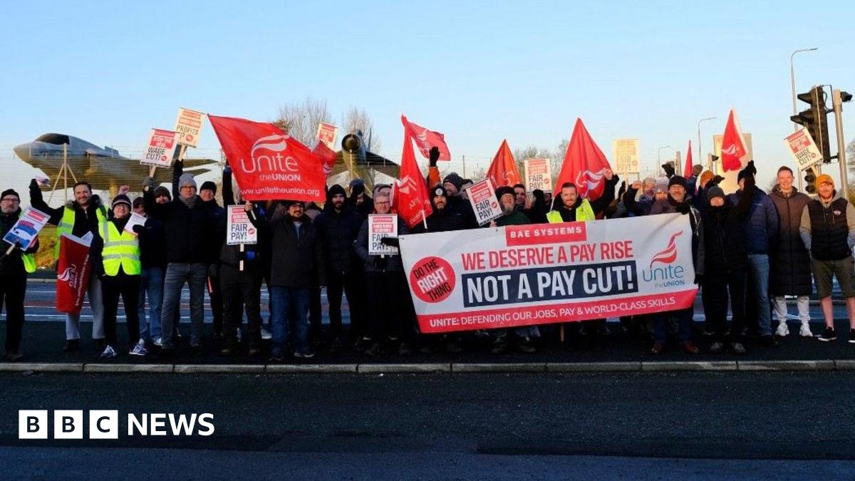 A small group of protesters holding flags and placards pushing for a pay rise. They are stood at the side of a road and dressed warmly in puffer jackets, jeans and woolly hats