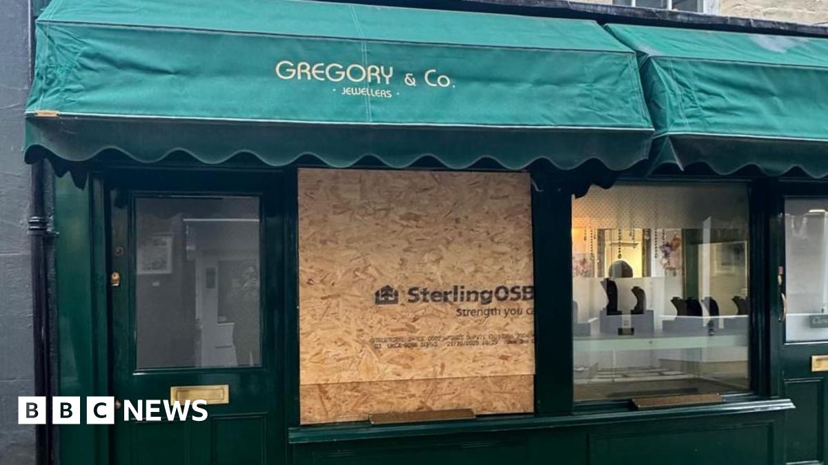 General street view of the boarded up shop front of jewelry store Gregory & Co. The window frames are painted green and there is a green marquis with the name written on it in gold letters. The large left-hand side window has been replaced by an OSB board which has 'Sterling OSB' written on it in bold black letters.