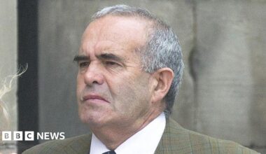 A man with short grey hair and black eyebrows pictured outside. He is wearing a green checked jacket and a shirt and tie