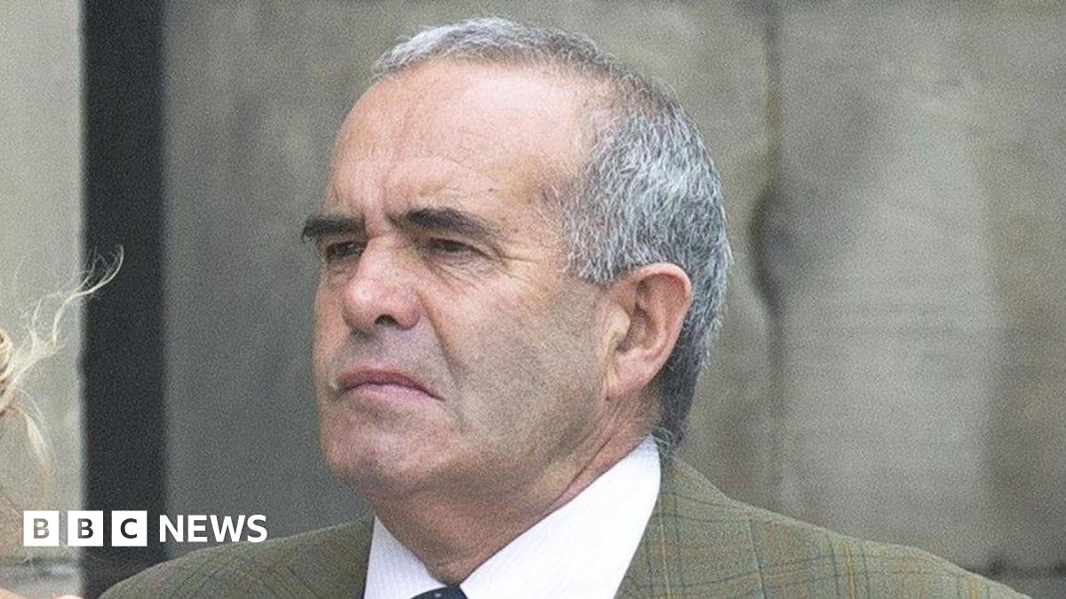 A man with short grey hair and black eyebrows pictured outside. He is wearing a green checked jacket and a shirt and tie