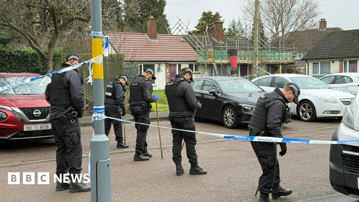 Four police officers in uniform in a line holding sticks looking for evidence as they move along a road on a residential street