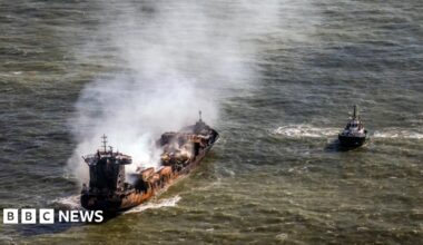 Smoke billows from a rust-coloured tanker ship floating in the sea. A tugboat floats nearby.