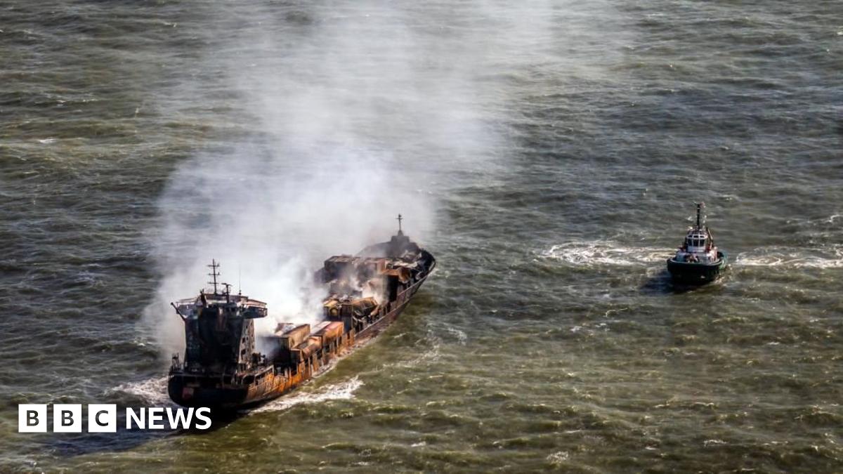 Smoke billows from a rust-coloured tanker ship floating in the sea. A tugboat floats nearby.