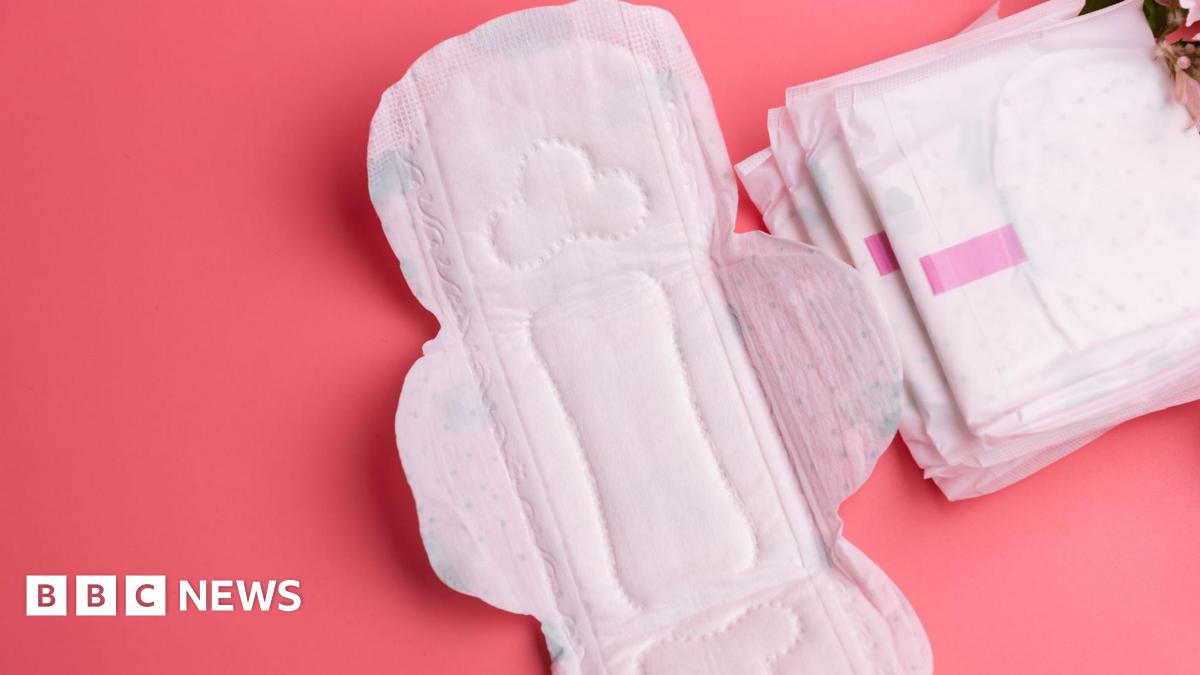 A white sanitary pad lies open with a pile of sanitary pads next to it, against a pink background