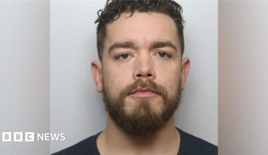 A mugshot of a man with short dark hair, a dark beard and a black shirt. He is looking directly into the camera.