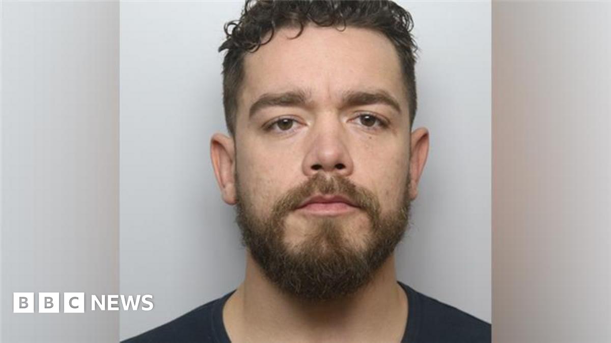 A mugshot of a man with short dark hair, a dark beard and a black shirt. He is looking directly into the camera.