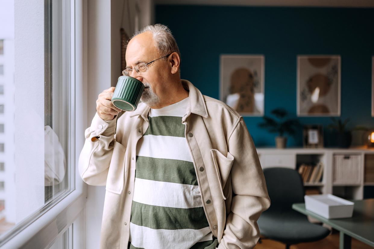 Thoughtful senior man stands by the window holding a coffee cup, enjoying a quiet moment and gazing outside in the morning light