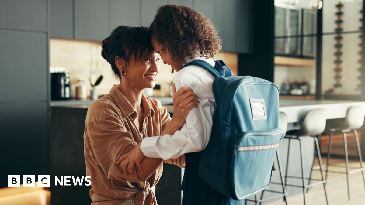 A parent kneels in a modern kitchen, holding and comforting a school‑aged child who is wearing a uniform and a large backpack, as they share a close and supportive moment.