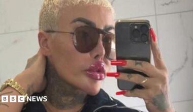 Jordan Parke seen with closely cropped bleached blonde hair takes a mirror selfie while holding a smartphone. They are wearing large dark sunglasses, a black zip-up top and a gold watch. Long, bright red manicured nails are visible as they hold the phone, and tattoos can be seen on their neck and hand. The background appears to be a tiled bathroom wall.