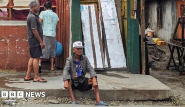 A damaged building with a partly collapsed roof and missing wall panels stands beside a roadside. Two people are standing in front of the structure, while another person sits on the pavement nearby.