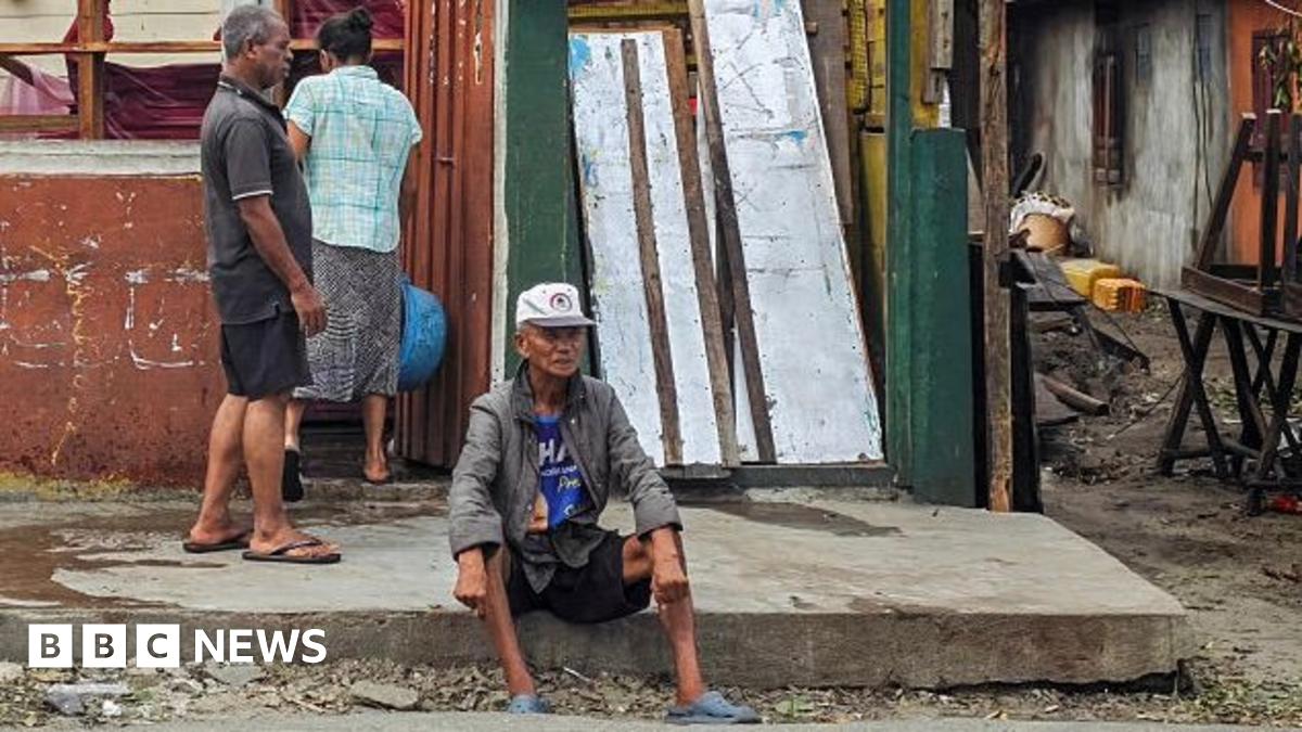 A damaged building with a partly collapsed roof and missing wall panels stands beside a roadside. Two people are standing in front of the structure, while another person sits on the pavement nearby.