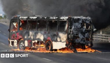 An armed Mexican police officer looks on while a bus burns on a highway in the background