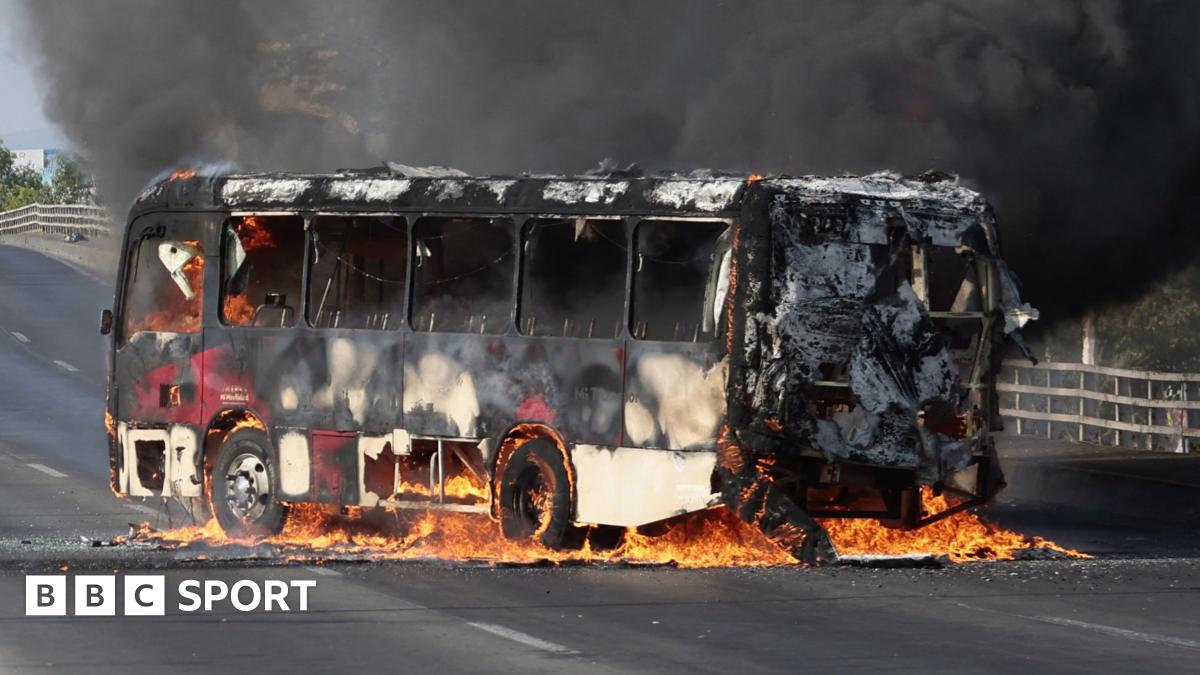 An armed Mexican police officer looks on while a bus burns on a highway in the background