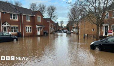 Two black cars in flood water near terrace brown homes. There is a 'For Sale' sign outside one of the homes.