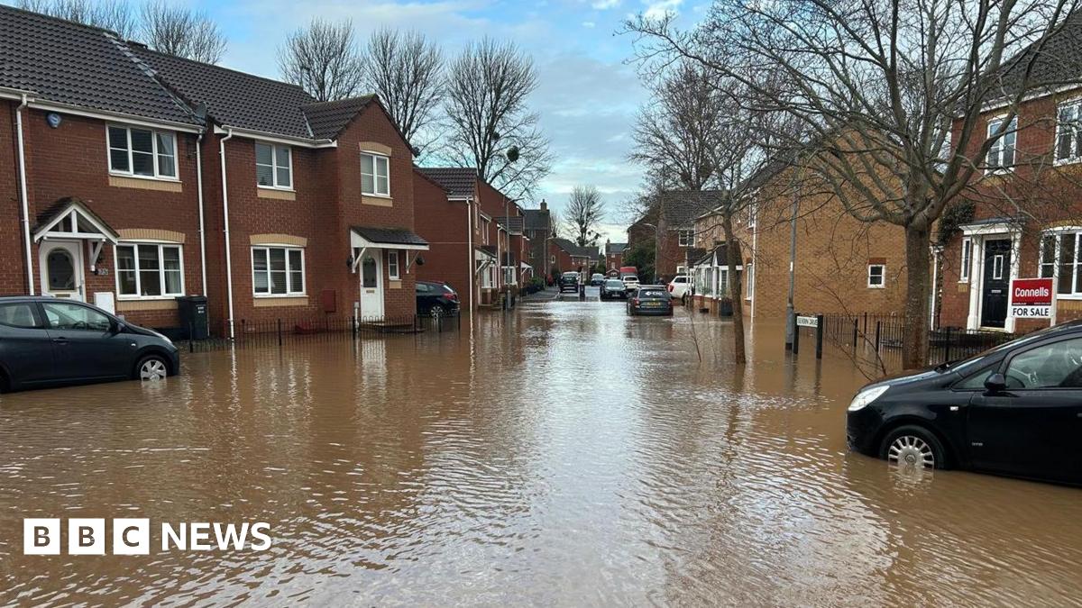 Two black cars in flood water near terrace brown homes. There is a 'For Sale' sign outside one of the homes.