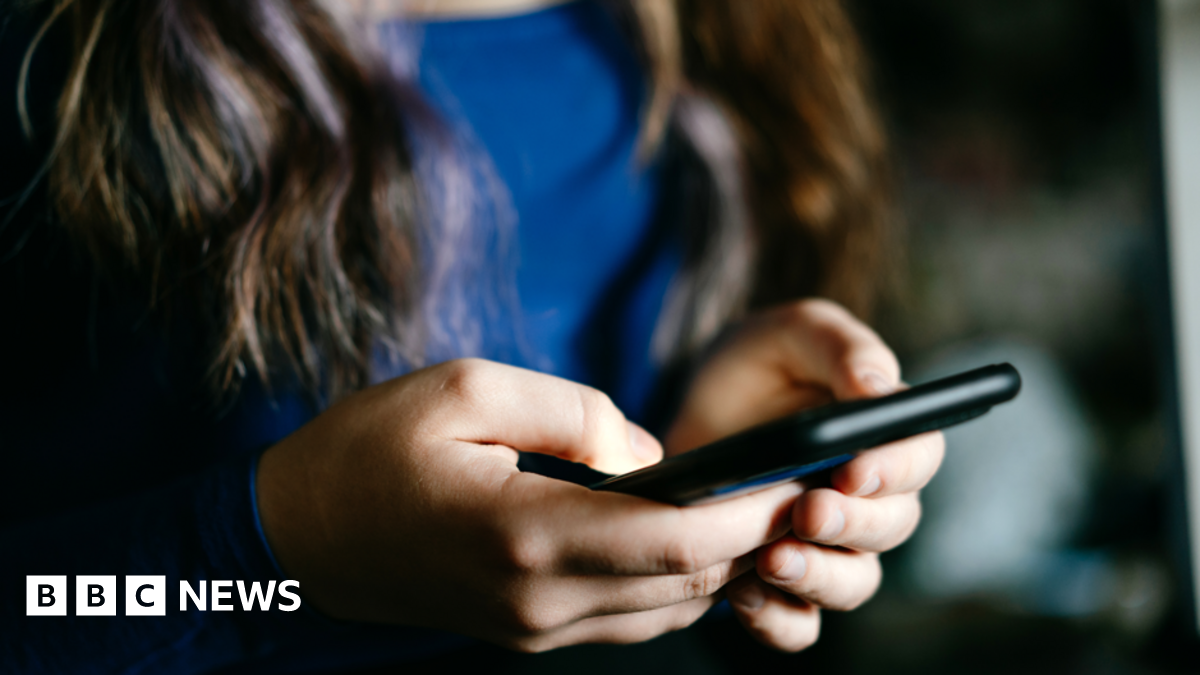 A close up stock image of an anonymous girl with long brown hair and wearing a blue top, holding a black smart phone in front of her.