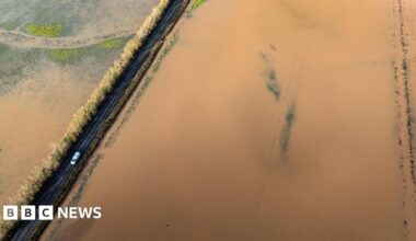 An aerial shot showing a car driving between flooded fields near the River Tone in Somerset.  The fields are almost completely invisible under brown floodwater