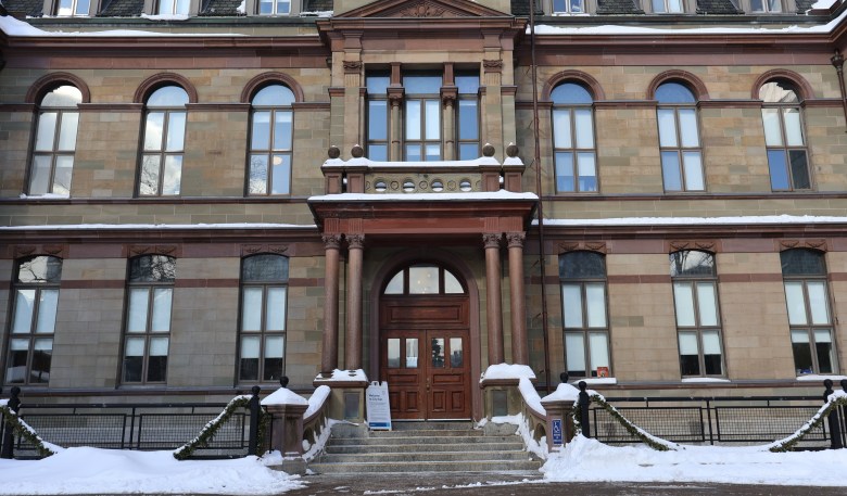 A closeup of a historic three-storey stone building. It has a wooden door at its main entrance and there's snow in the front on either side of the front staircase.