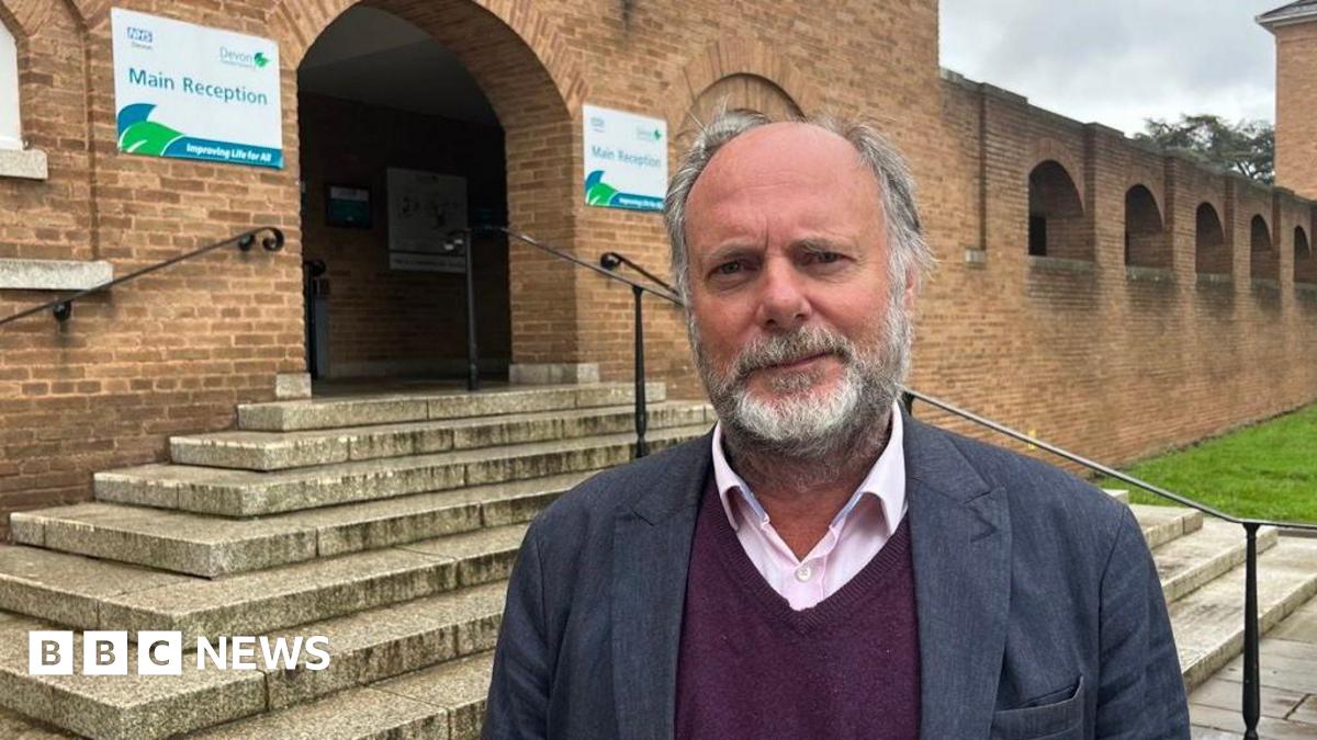 Julian Brazil is wearing a light pink shirt and a maroon V-neck sweater under a dark blue suit jacket and is standing in front of steps at the main entrance to the red brick-fronted County Hall in Exeter.