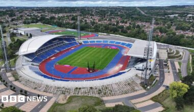 An aerial view of the stadium, which dominates the photo. Covered stands by the blue athletics track are on the left and right of the image. Trees and houses are in the background.
