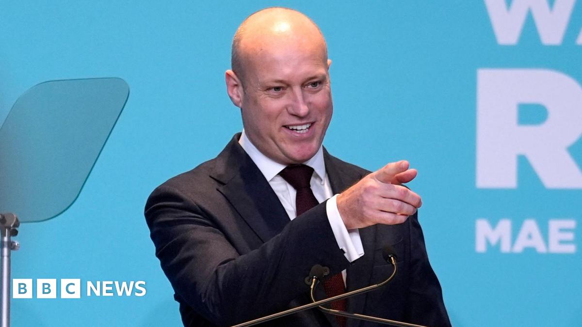 Dan Thomas on stage smiling behind a lectern with a Reform UK banner on it at a rally in Newport. He is pointing at someone in the audience.