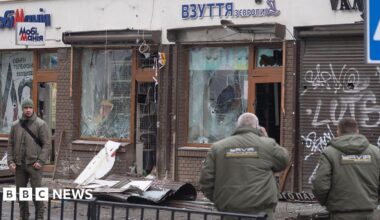 Three people stand in front of a building with broken windows. Debris is strewn on the pavement in front of it.