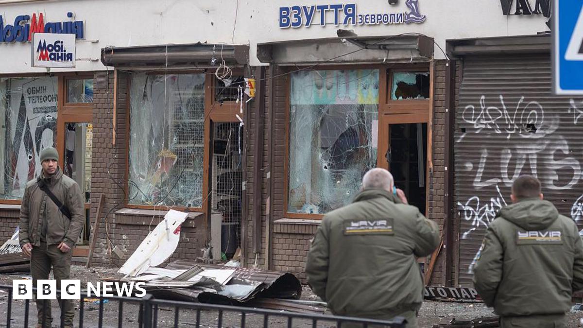 Three people stand in front of a building with broken windows. Debris is strewn on the pavement in front of it.
