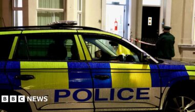 A police car outside a cream house. The door is open and an officer standing outside. The car is green white and blue.