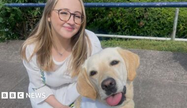 Megan Stephenson is crouching down with her assistance dog. She is smiling and has long blonde hair and is wearing glasses. She is wearing a white dress. Her dog is in the foreground of the photo.